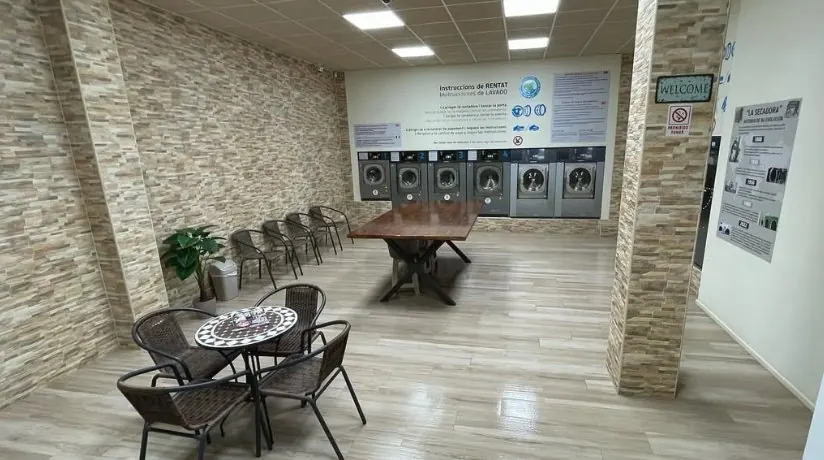 Rincón de espera con mesa redonda y sillas, y la mesa grande de doblar delante de las lavadoras
