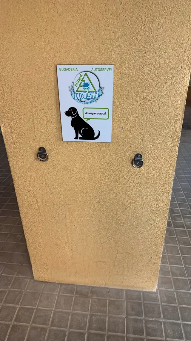 Pilar junto a la puerta con un cartel de un perro sentado que dice «Jo espero aquí!» y dos anillas metálicas para atar la correa